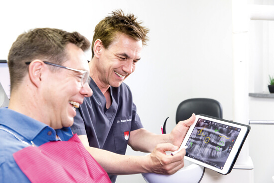 Implantologie: Dr. Heckel zeigt einem Patienten am Tablet den Plan für die künftigen Implantate