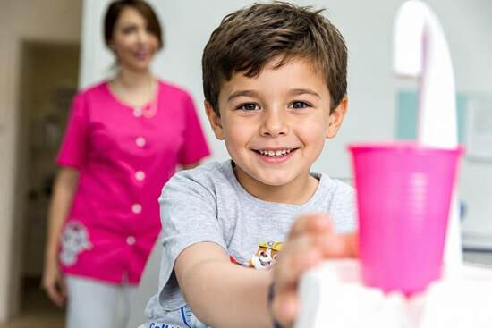 Kinderzahnheilkunde: ein lächelndes Kind steht vor einem rosa farbenen Becher mit Mundspülung